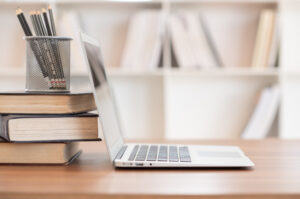 Photo of a stack of books, pencils in a cup and a laptop, all sitting on a desk in front of a bookcase